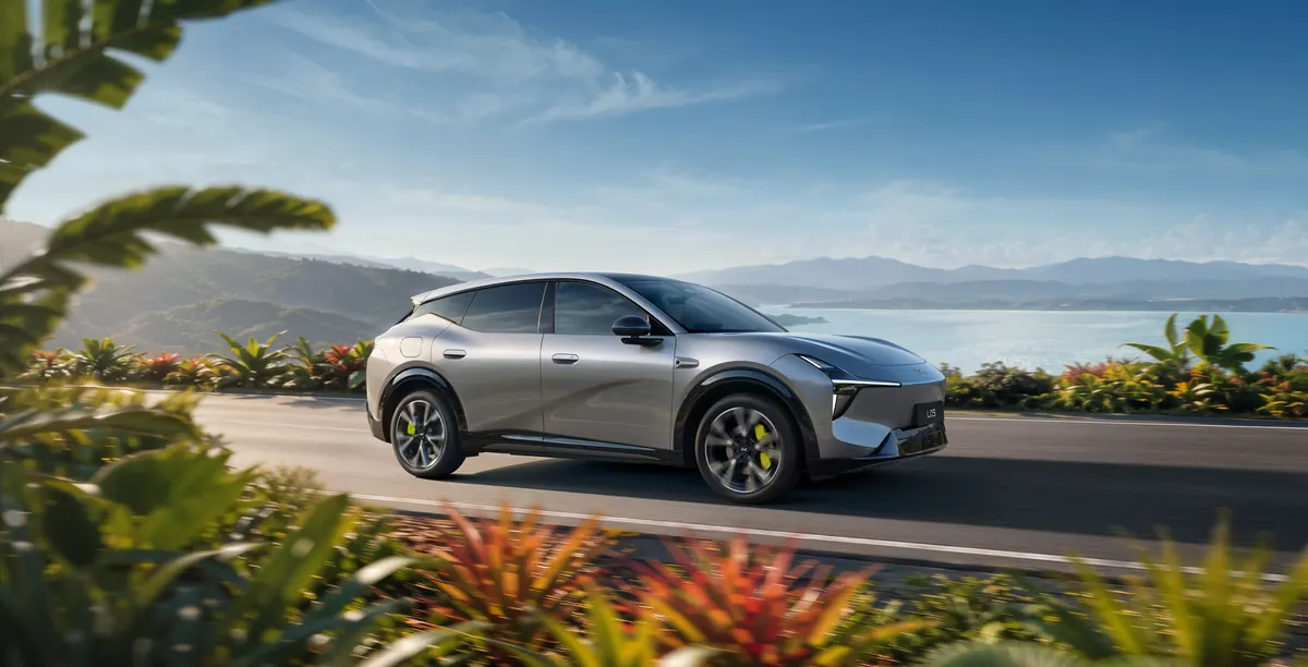 A silver MONA L05 SUV driving along a coast road, with flowering plants in the foreground and a bay and hills behind.