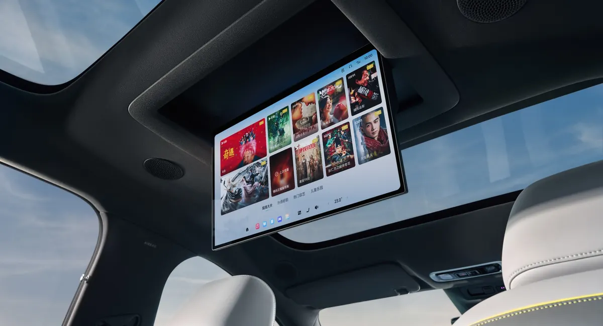 The MONA L05’s 17.3-inch rear screen folded down from the roof, showing a menu of films, with light grey seats and a glass roof around it.