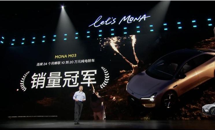 He Xiaopeng on stage beside the MONA M03, which XPeng calls the top-selling electric sedan at RMB100,000–200,000 for 24 months running. XPeng's broadcast, 11:09 UTC.