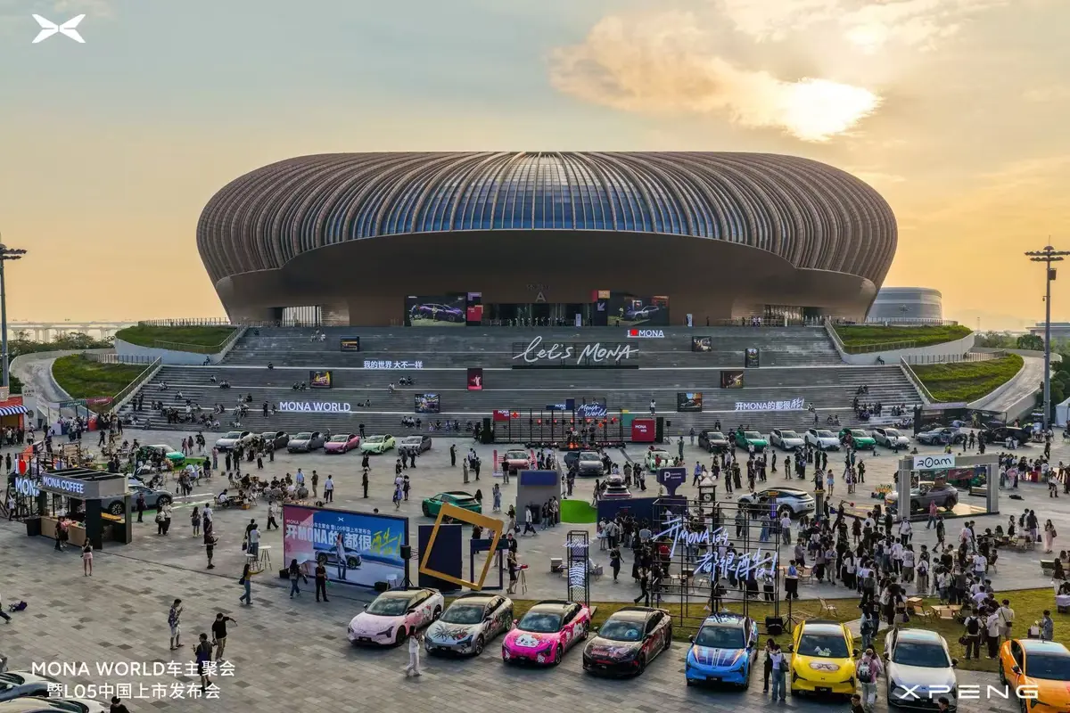 The Guangzhou stadium at dusk, seen from above, with MONA owners and their cars gathered on the square in front of it under Let’s MONA signs.